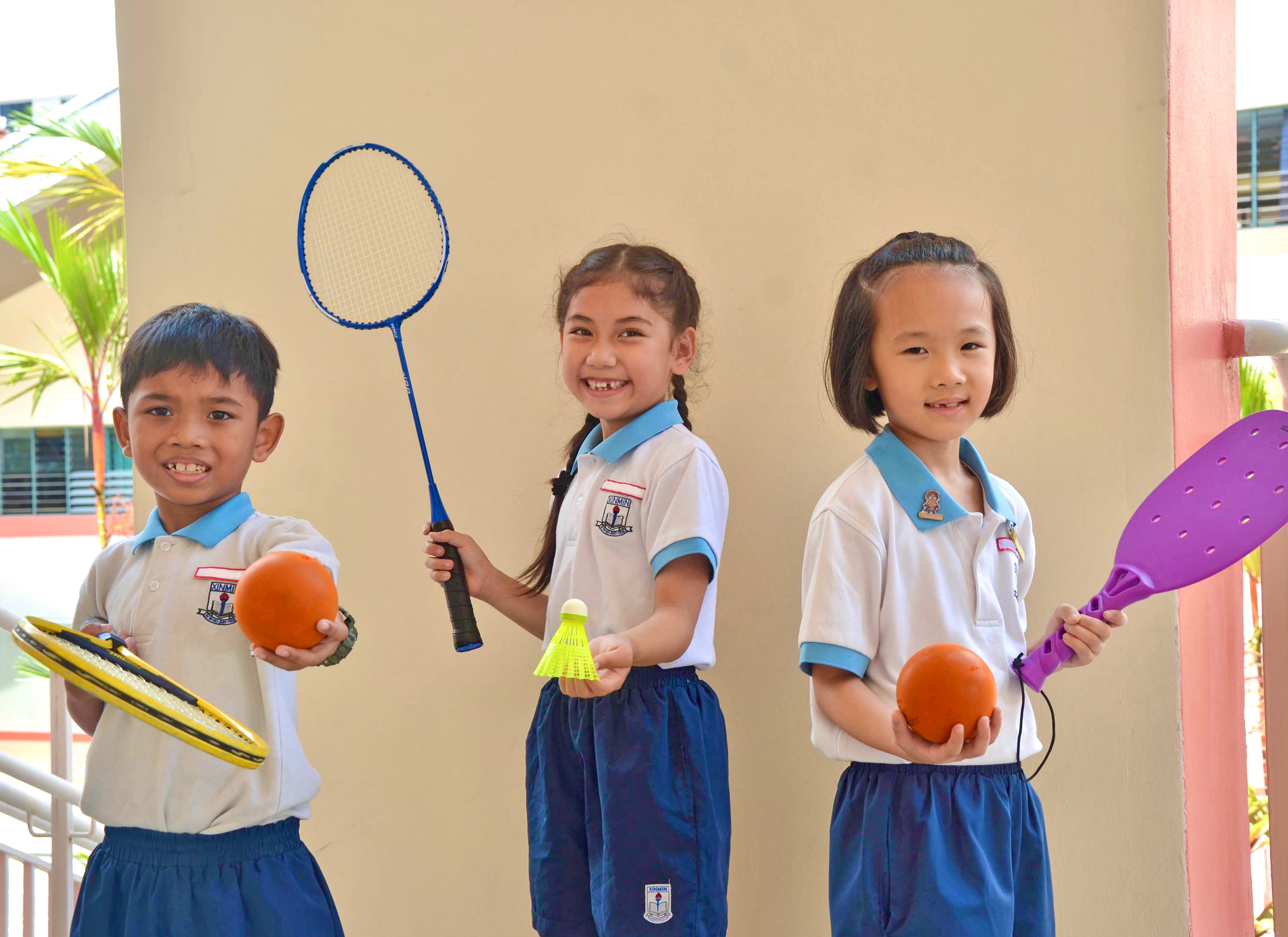 Three smiling children in school uniforms hold sports equipment including badminton rackets, a shuttlecock, and orange balls.